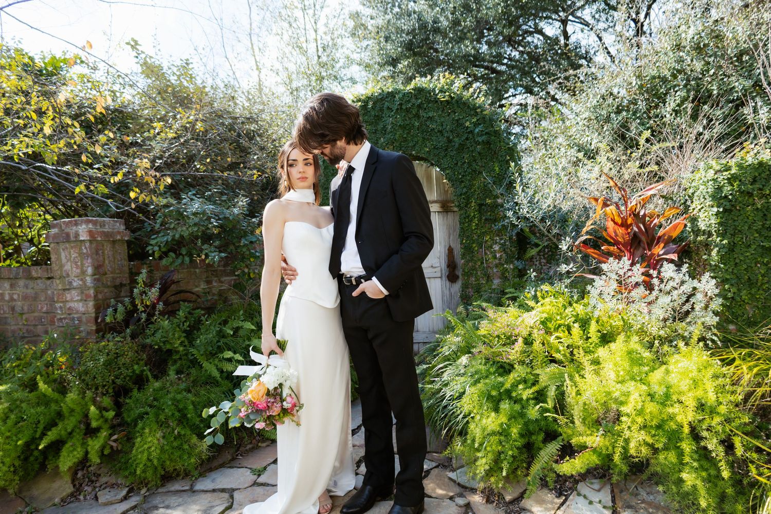 A couple in wedding attire stands together in a lush garden setting with greenery and flowers surrounding them.