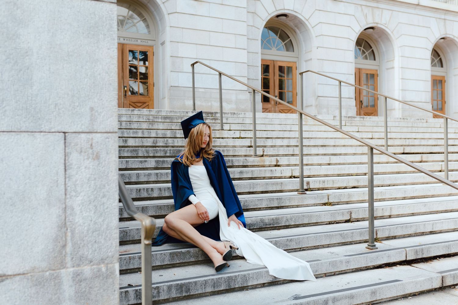 UC Berkeley graduate wears a white dress and poses elegantly for her graduation photoshoot on the marble steps of Doe Library.