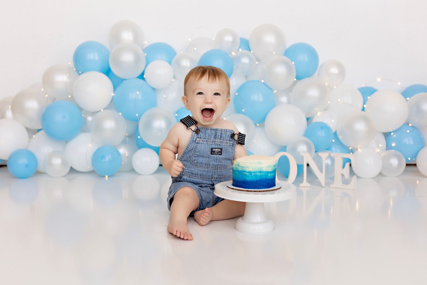 Baby in denim overalls celebrates first birthday with blue and white balloon backdrop and smash cake setup.