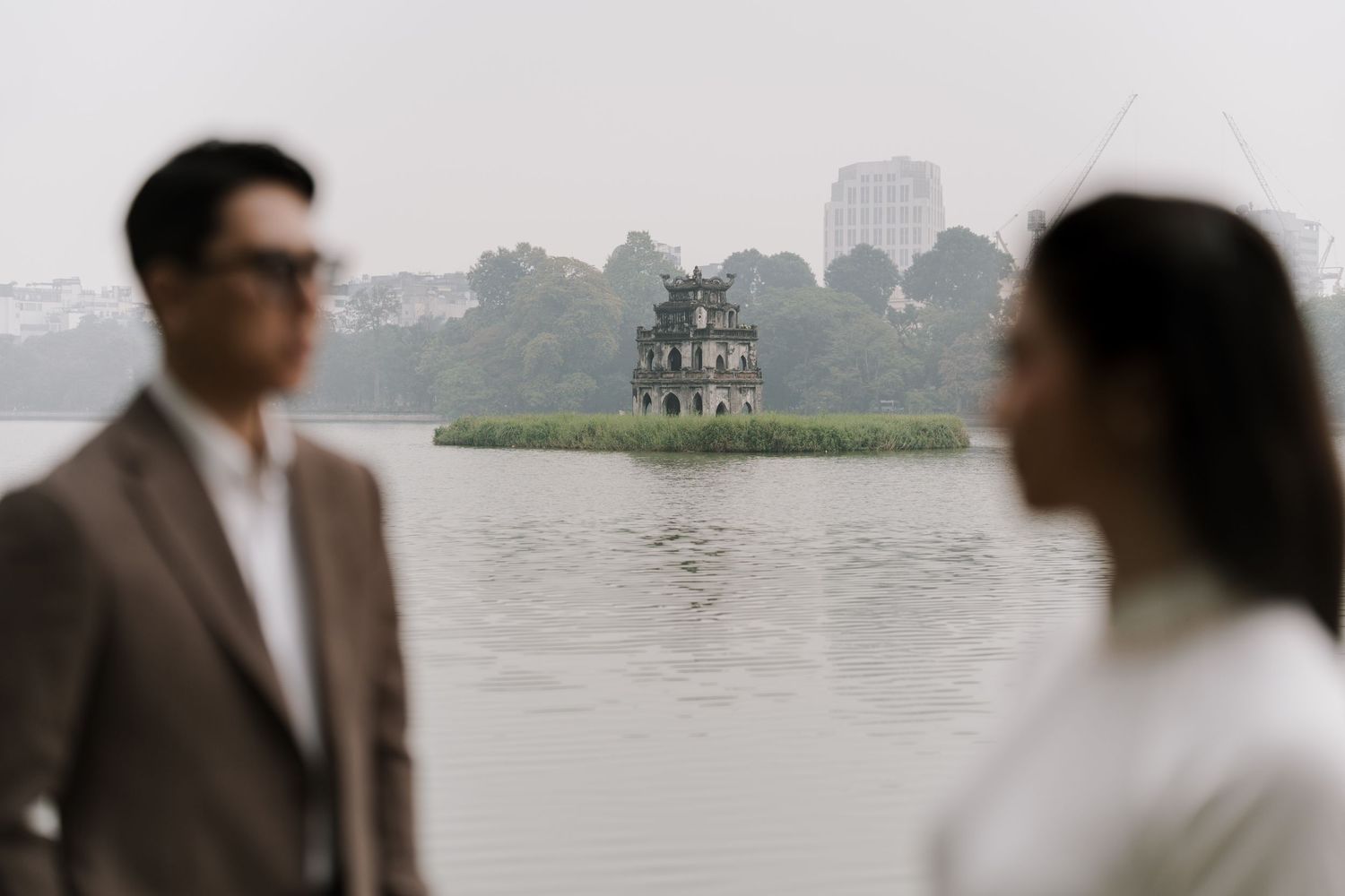 Couple standing near Hoan Kiem Lake with Turtle Tower visible in misty background, Hanoi Vietnam.