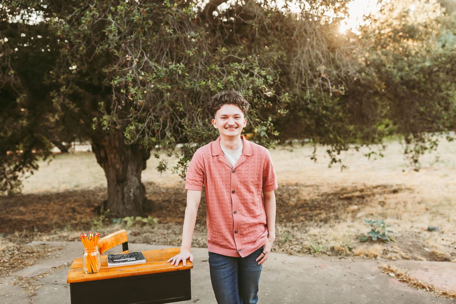 A person in a coral shirt stands in a sun-dappled outdoor setting with natural lighting and trees in the background.