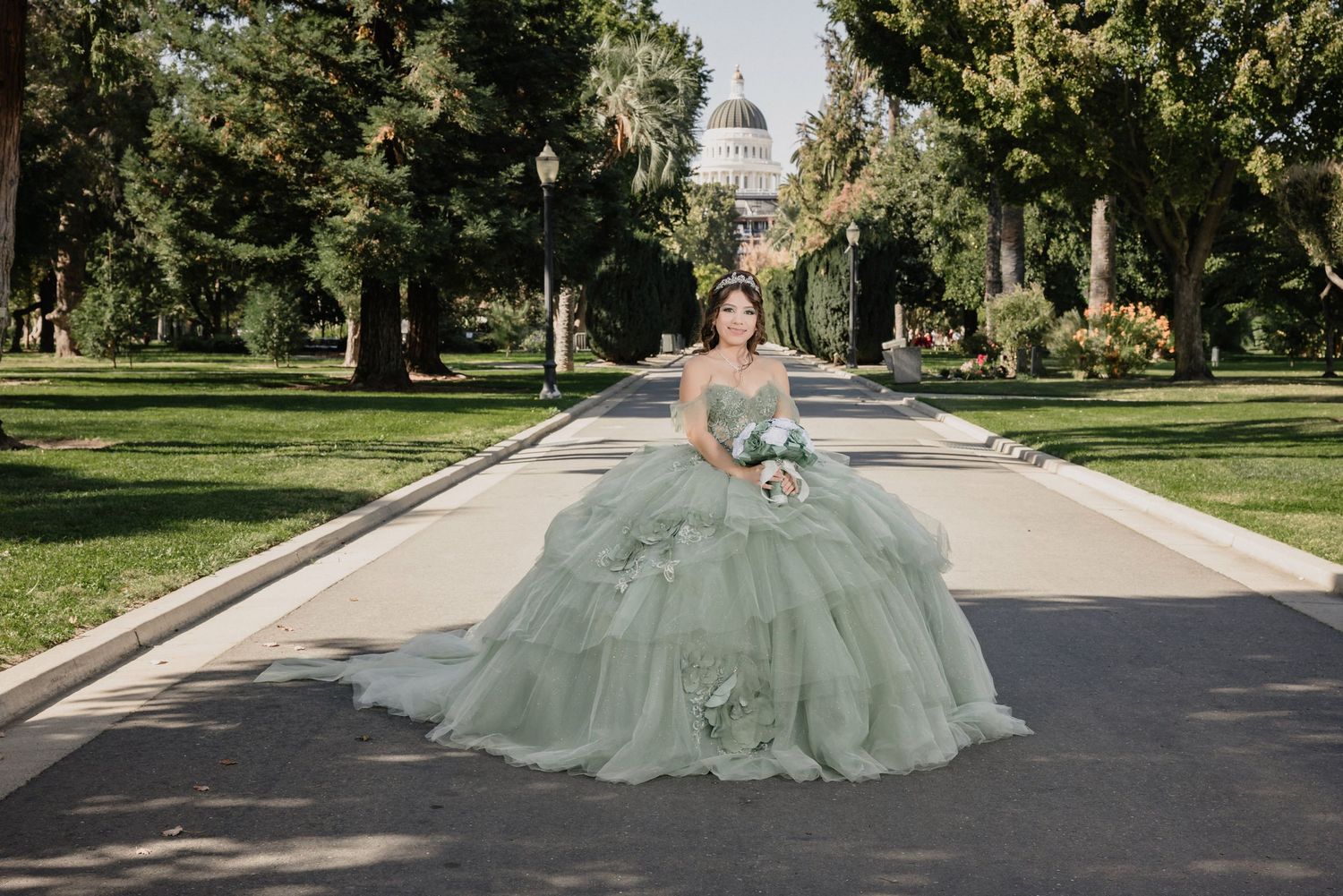 A light green dress flows elegantly on a tree-lined path with the State Capitol building visible in the background for Sacramento quince
