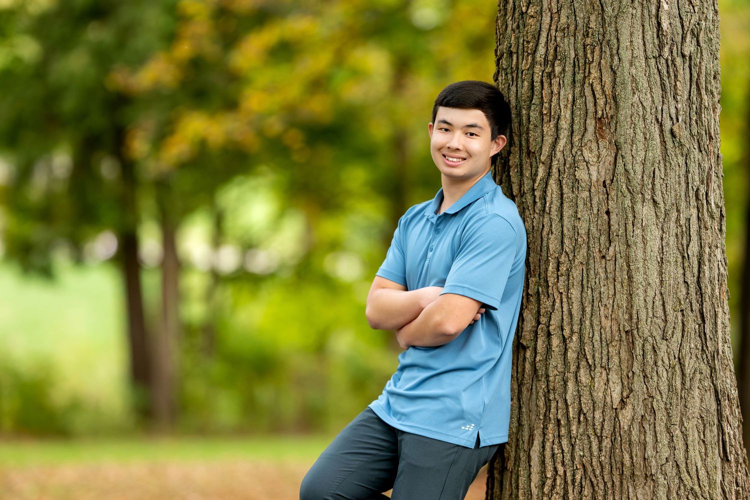 Smiling young man in blue polo shirt leaning against tree with arms crossed in green park.