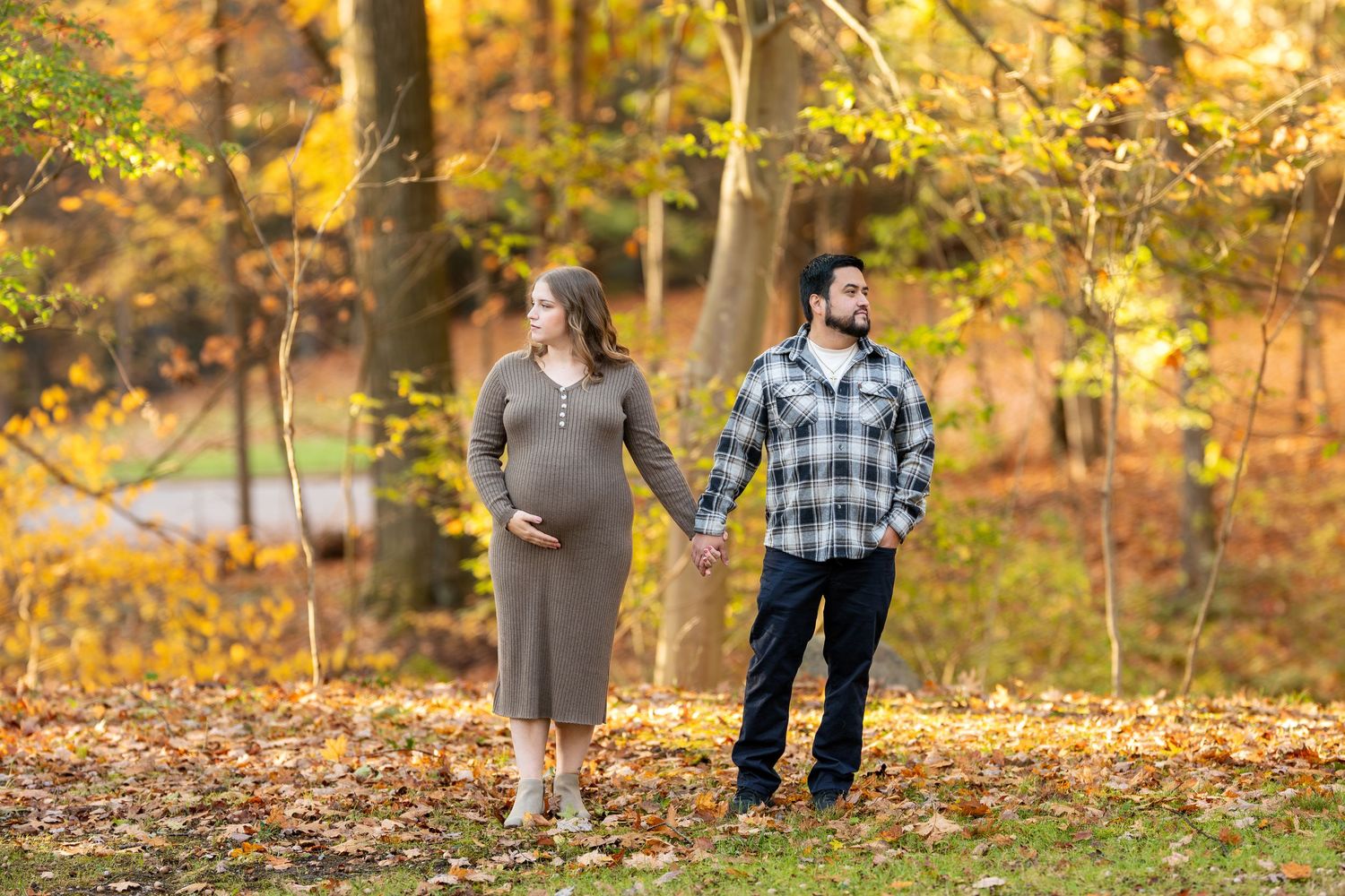 Pregnant couple holds hands while walking among golden autumn leaves in a park.