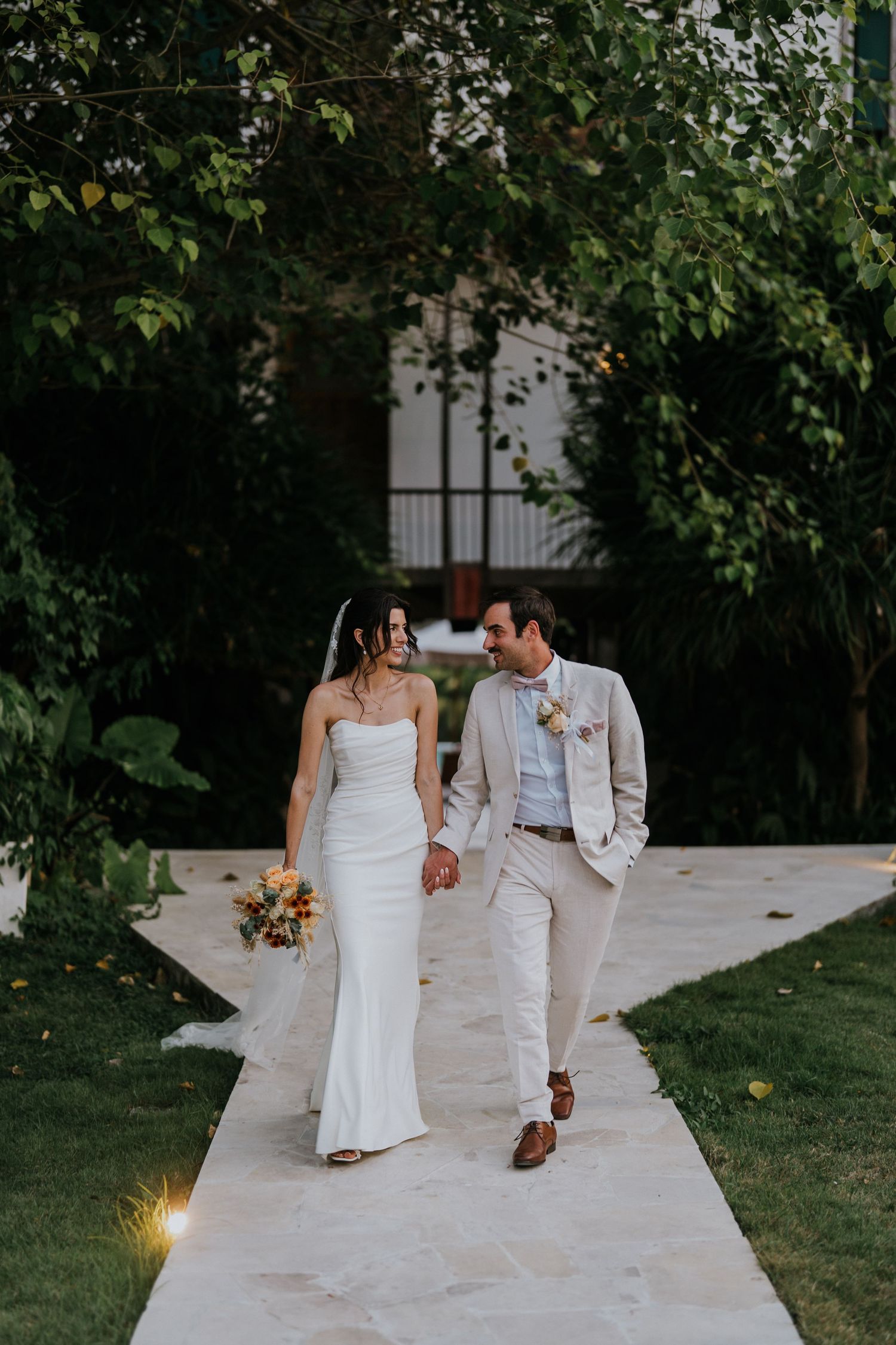 A couple walks hand in hand along a white pathway surrounded by greenery.