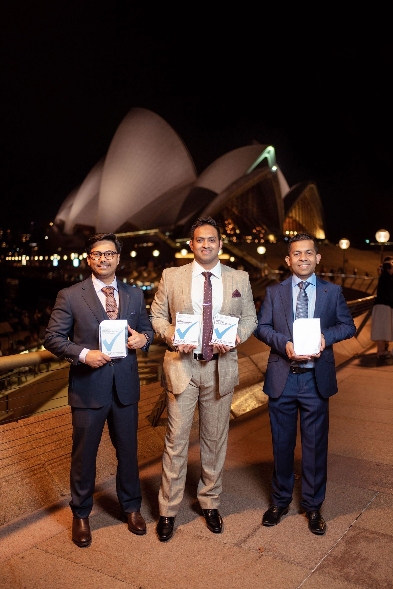Three people in suits pose with drinks at a nighttime rooftop venue overlooking city lights.
