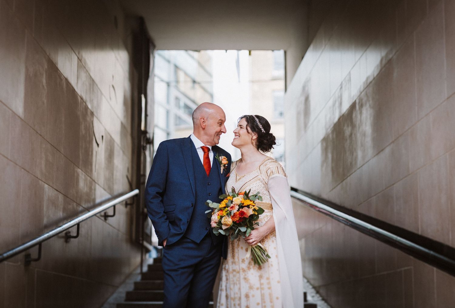 A couple wearing wedding attire share an intimate moment in a white stone hallway with handrails.