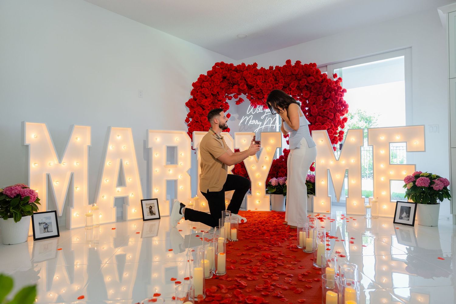 Illuminated "MARRY ME" letters with vibrant red rose decorations set up for a romantic indoor proposal setup.