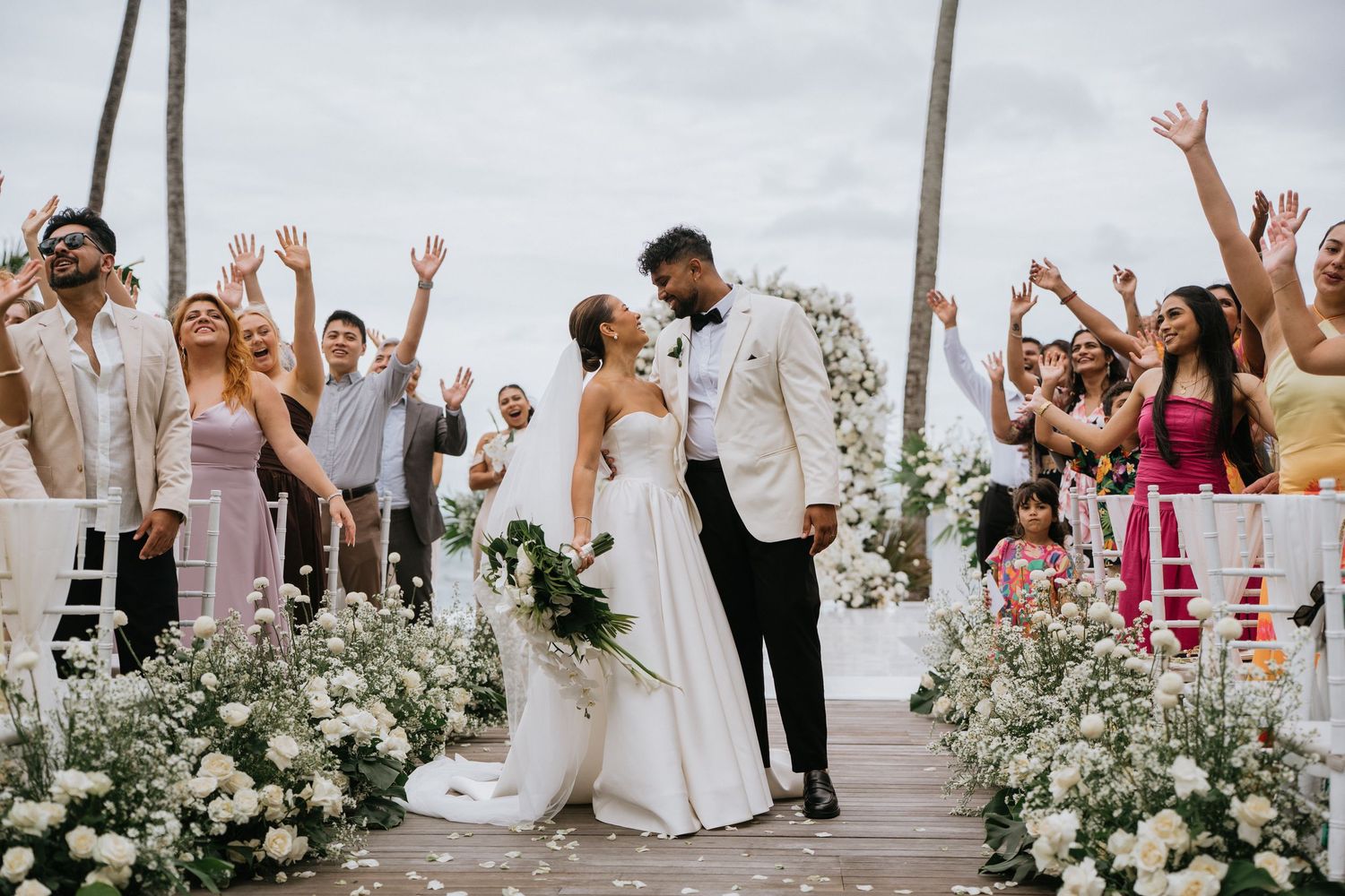 Joyful wedding celebration with guests dancing and cheering in an outdoor setting with white floral decorations.