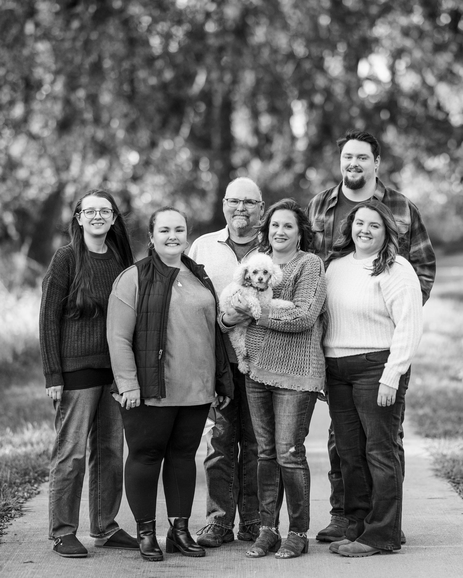Black and white outdoor portrait of a family group of six people and a small dog standing together on a path.