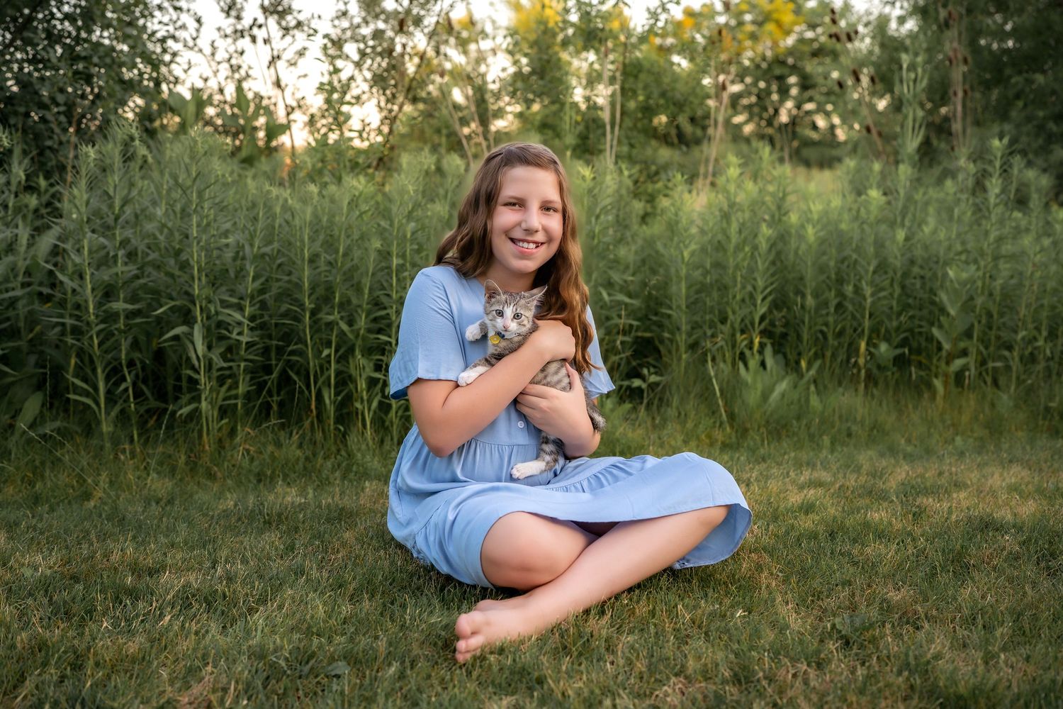 A person in a blue dress sits on grass holding a small grey kitten near tall prairie grass at sunset.