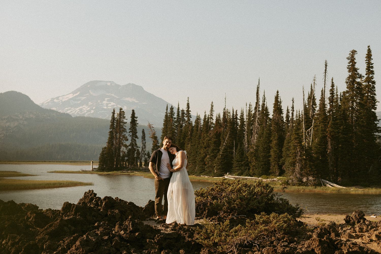 Couple embracing on volcanic rocks beside a calm lake with evergreen trees and a snow-capped mountain in the background.