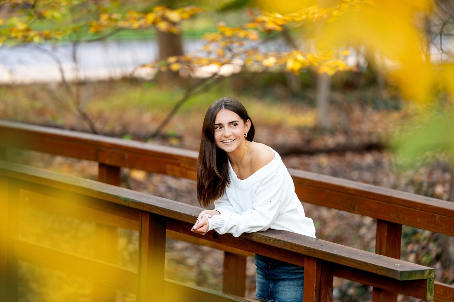 Smiling young woman with long dark hair in a white sweater leans on a wooden railing surrounded by golden autumn foliage.