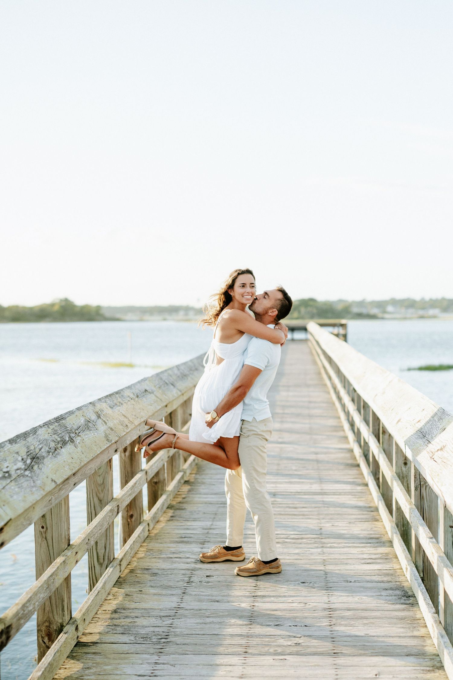 Couple in white outfits embracing on a wooden pier overlooking calm water during golden hour.