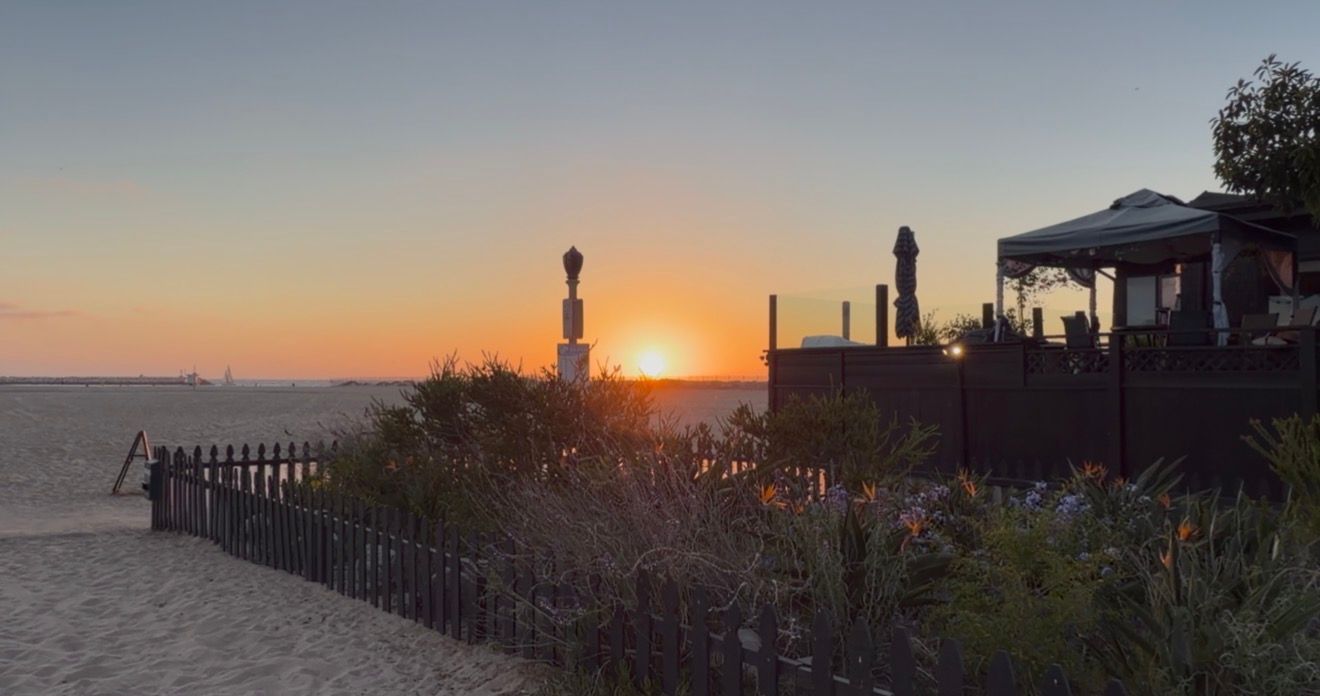 Stunning beach sunset with golden sky, sandy shore, wooden fence, tropical plants, and outdoor dining area silhouettes.
