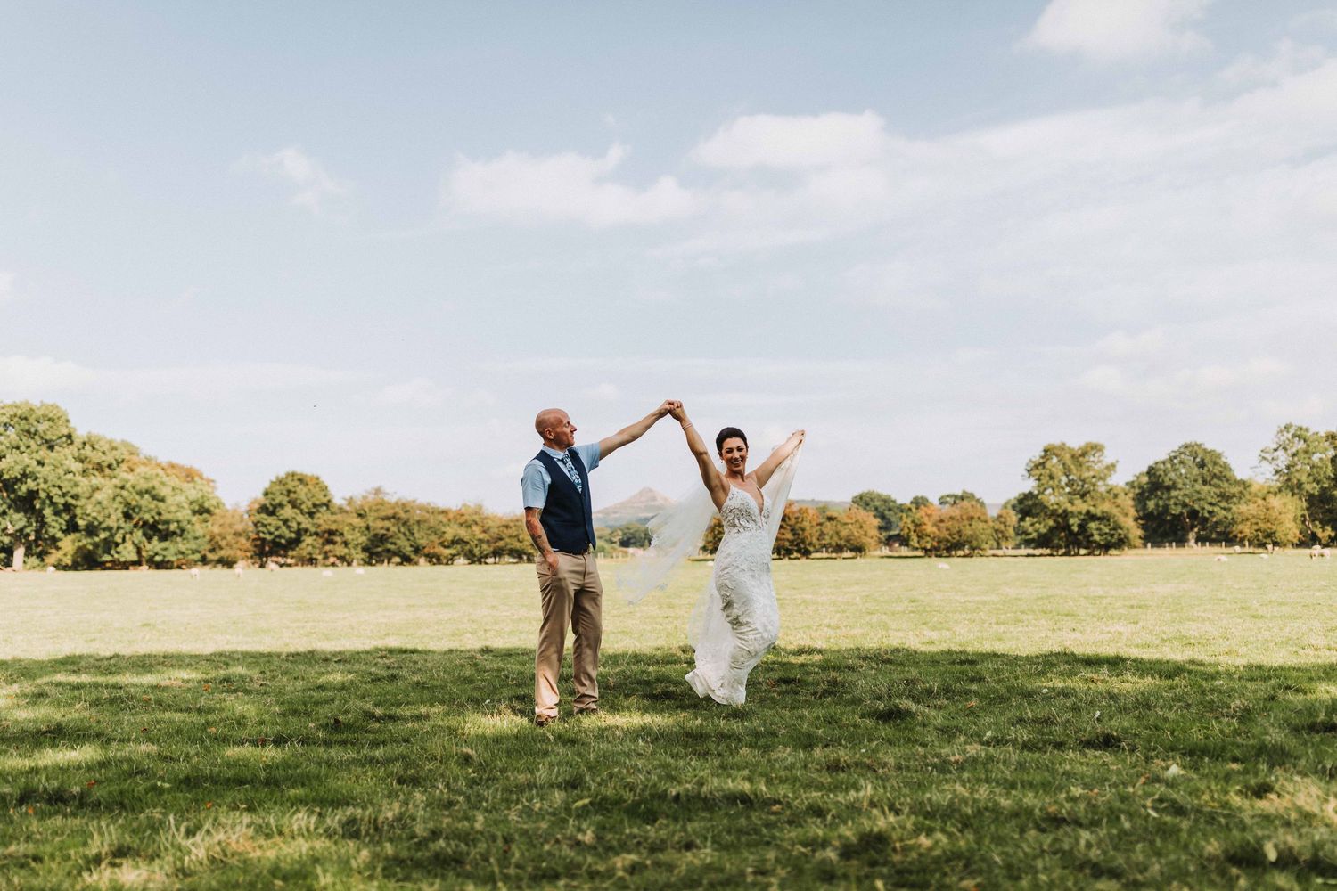 Bride and groom dancing in a sunny green field, with the bride's veil flowing in the wind on their wedding day.