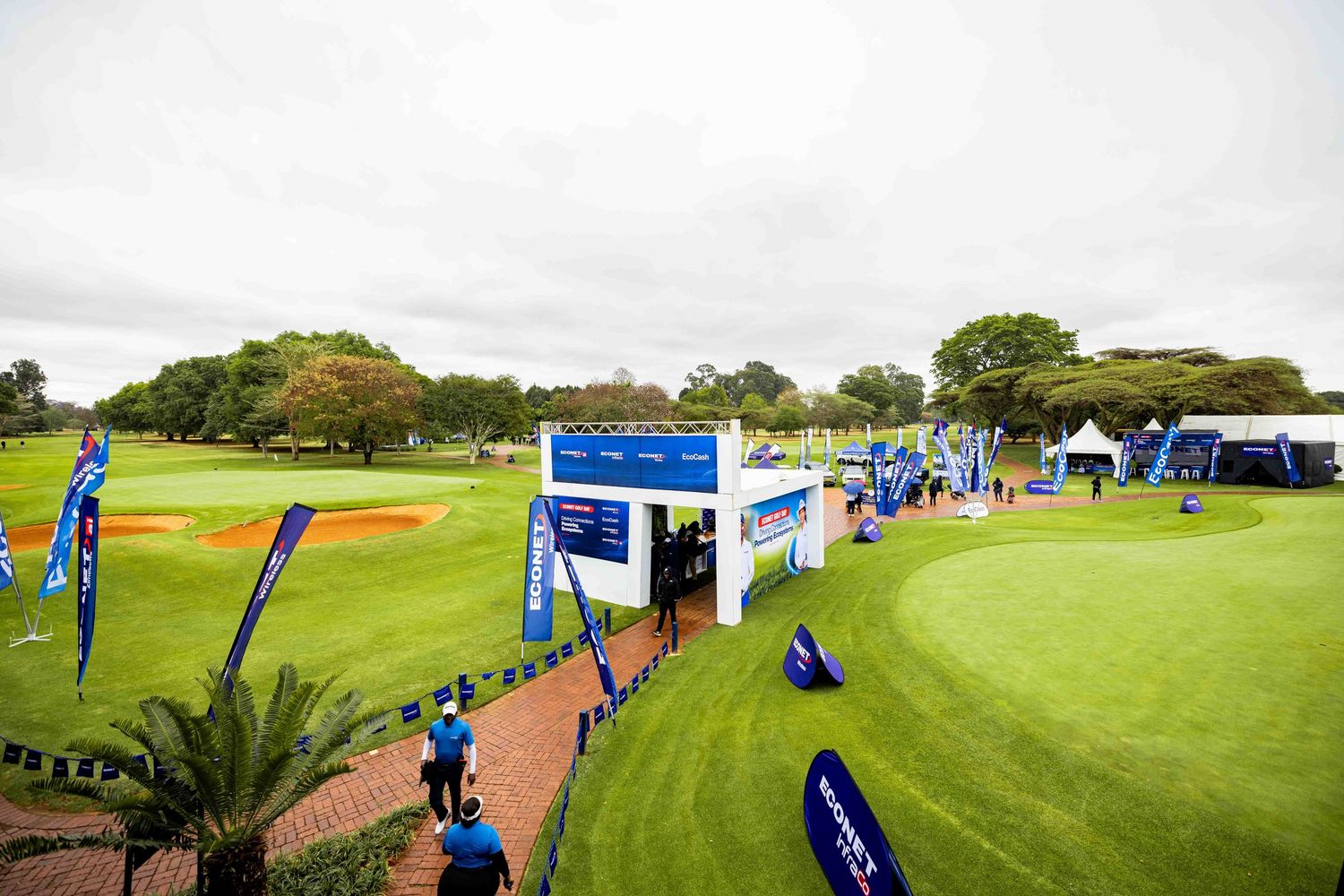 Group of caddies in matching blue shirts and white hats pose with golf bags before an Econet Golf Day banner on a sunny green course.