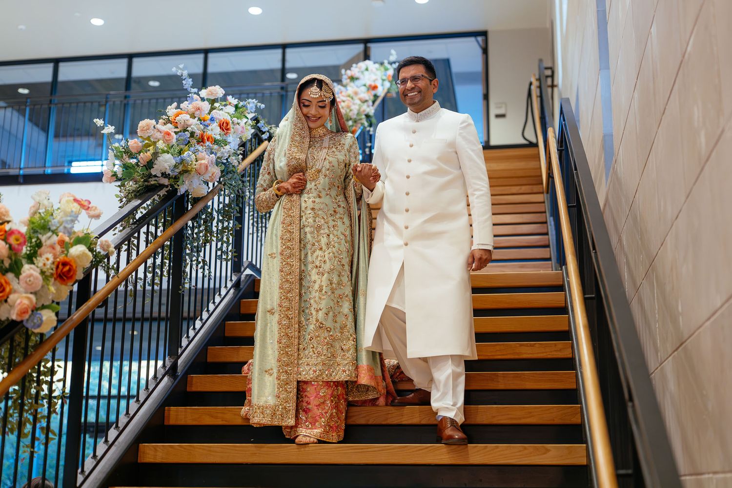 Bride in an ornate gold embroidered gown and groom in a white sherwani descending a wooden staircase decorated with floral arrangements.