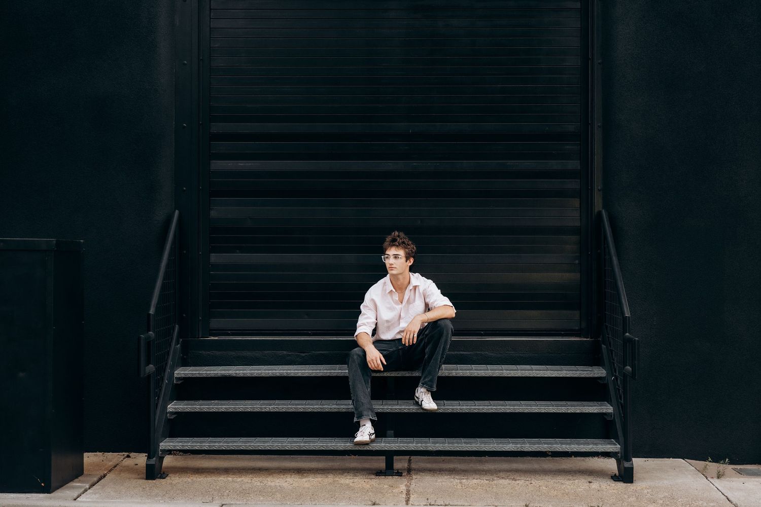 Young person sitting on black metal stairs against a dark wall, wearing white shirt and black jeans.