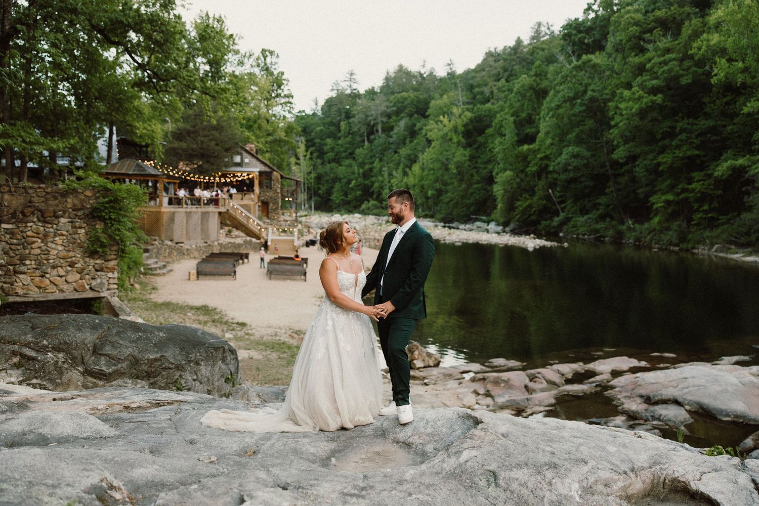 Bride and groom holding hands on rocky riverbank with lush forest and rustic venue in background.