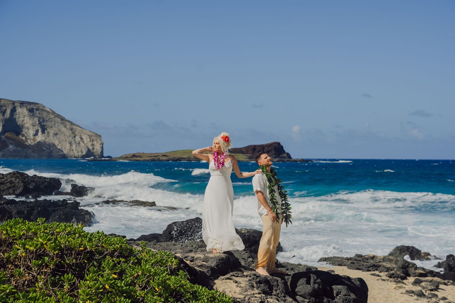 Black and white photo of a couple embracing on rocky Hawaii shoreline, wearing leis, with ocean waves crashing behind them.