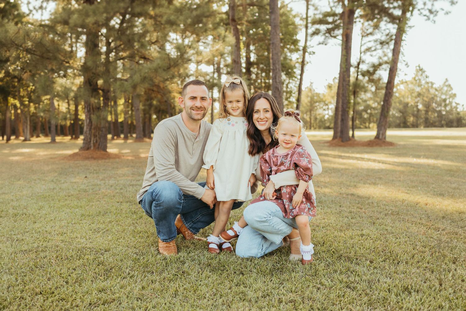 A happy family of four crouches together on green grass surrounded by tall pine trees in warm sunlight.