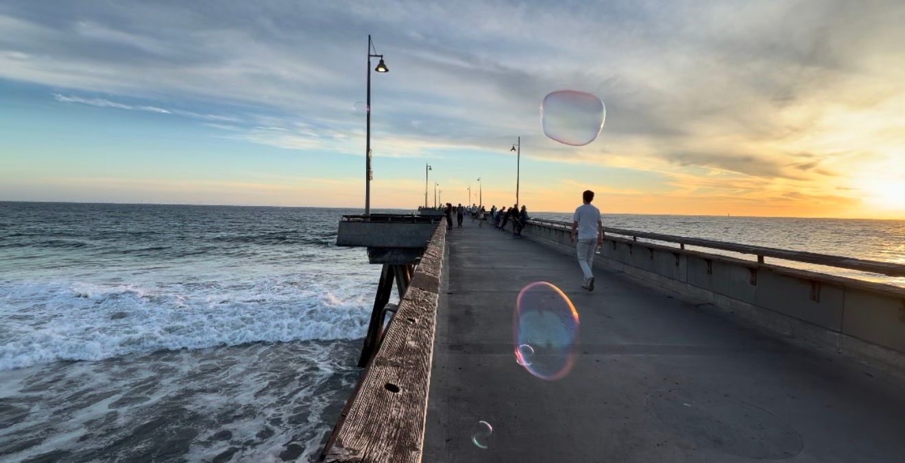 Person walking on a seaside pier at sunset with soap bubbles floating in the foreground and waves crashing below.