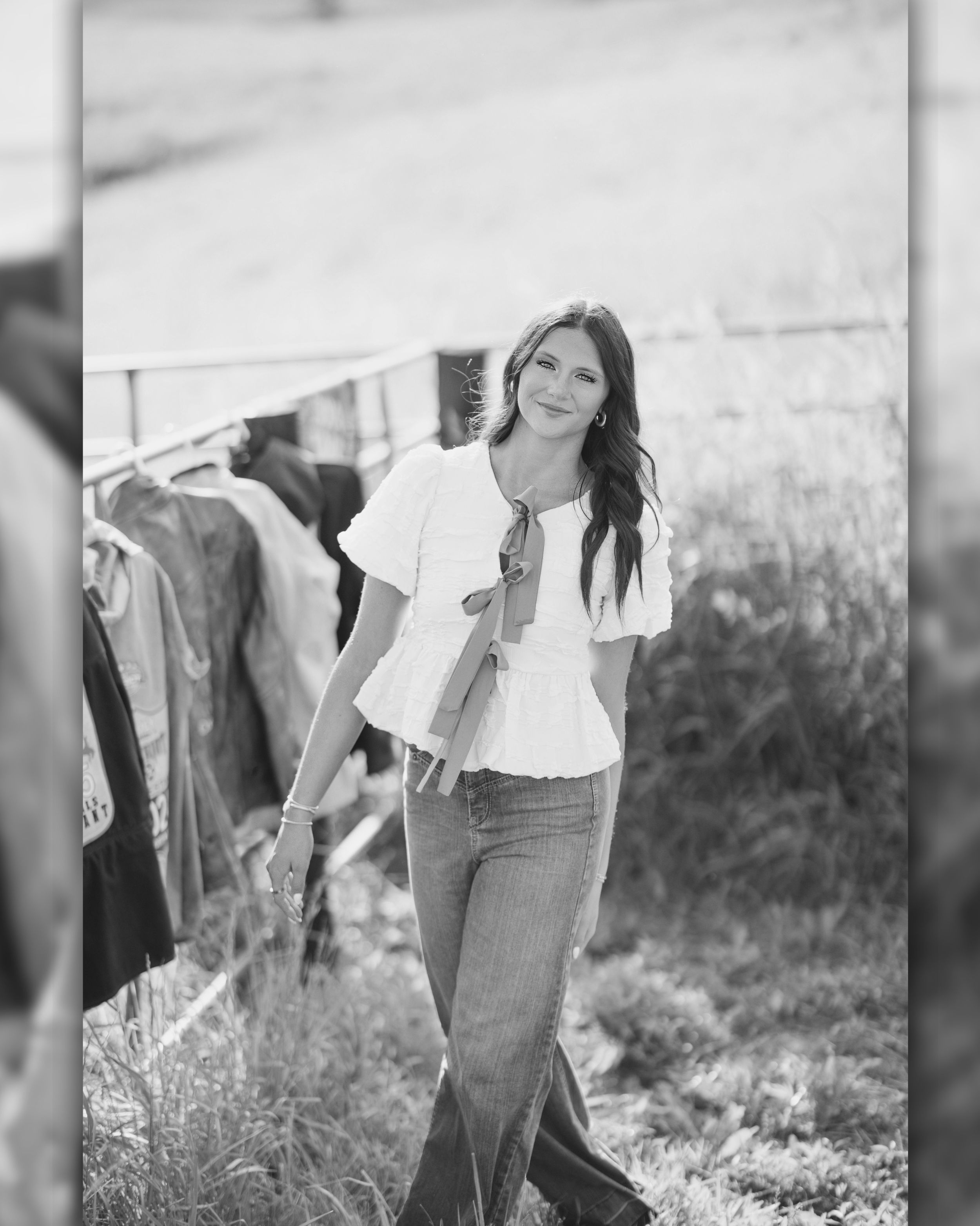 Woman in white blouse and jeans smiling outdoors near clothing rack in black and white photo.