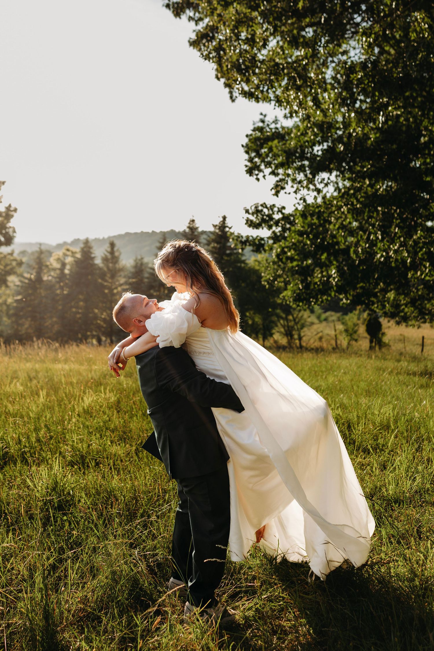 Groom lifting bride in white dress outdoors in sunny green field with trees, romantic wedding photo.