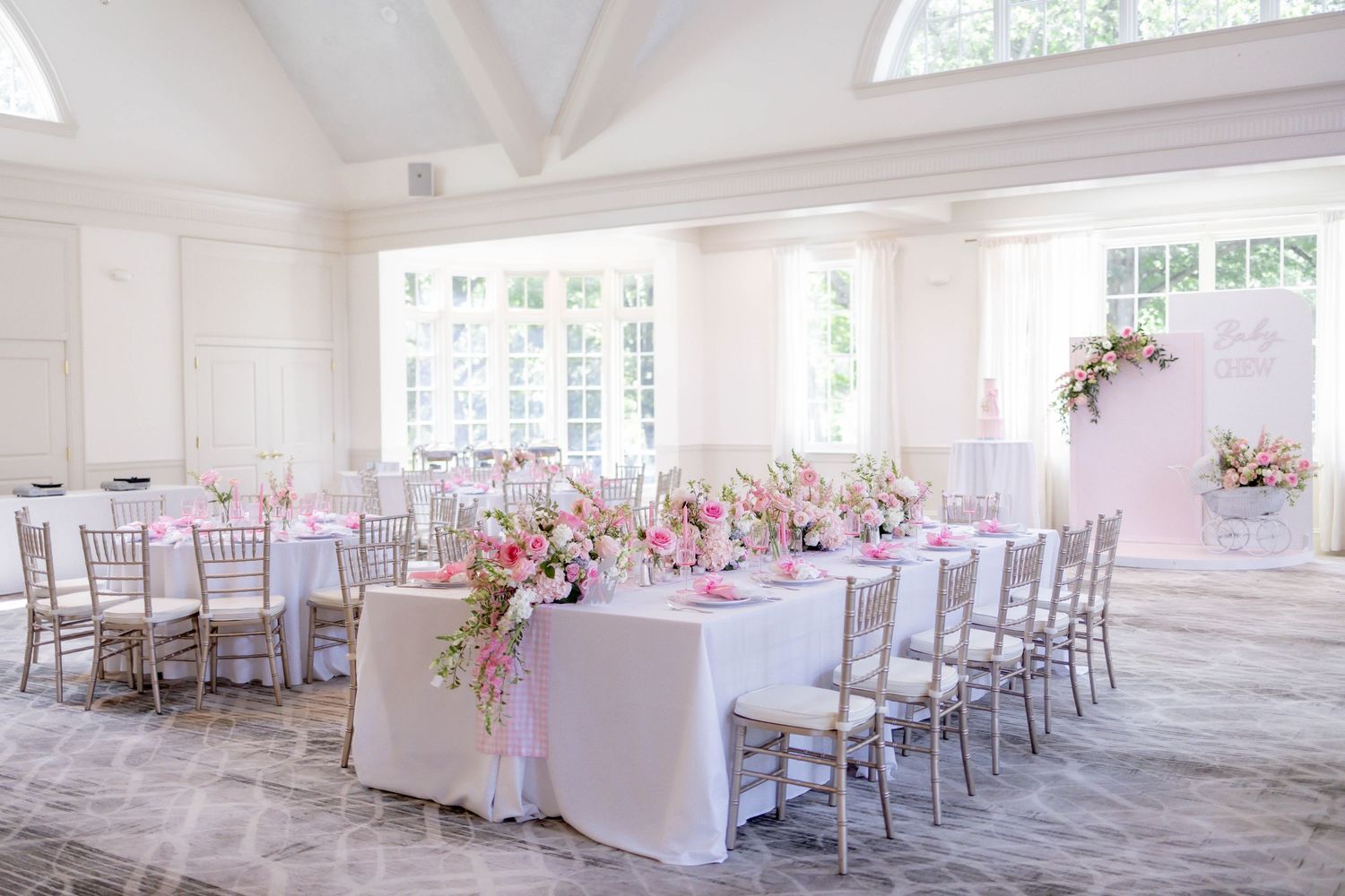 Elegant wedding reception hall with pink floral centerpieces, white tablecloths, and silver chiavari chairs in a bright venue.