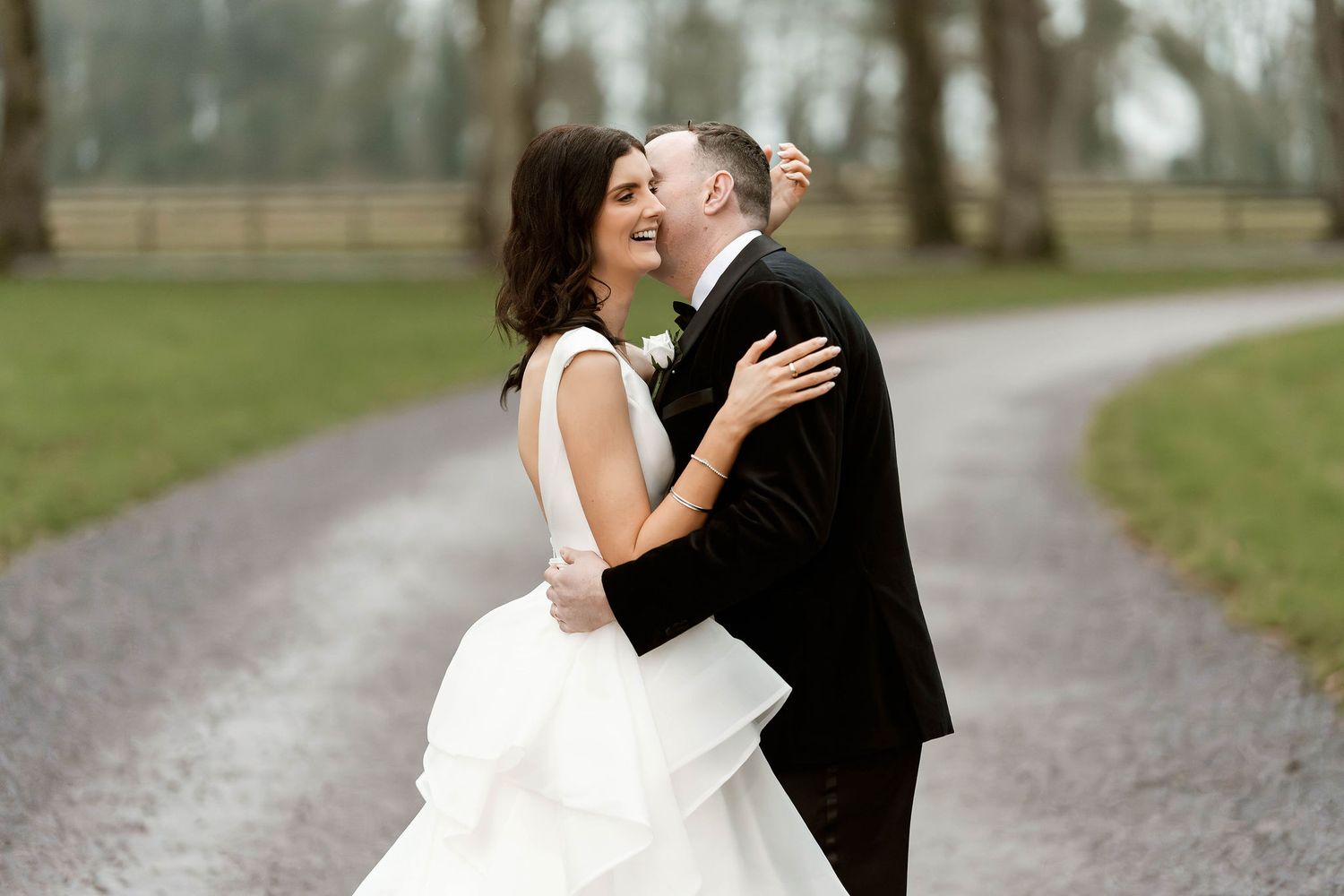 A joyful bride and groom embrace on a tree-lined path, laughing together on their wedding day.