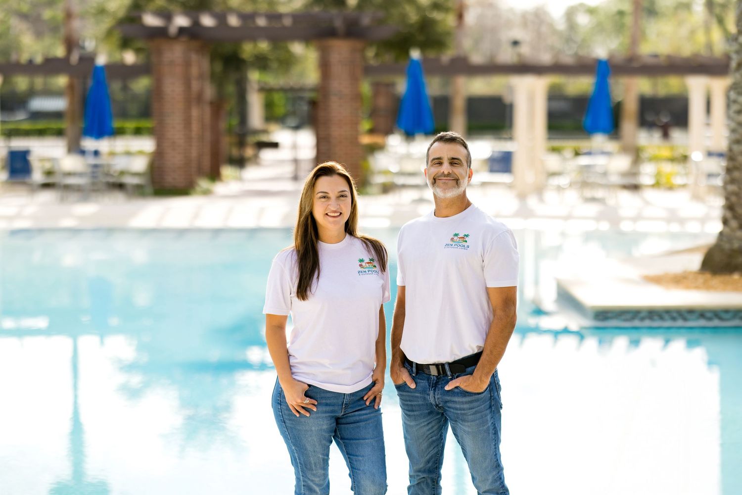 Two smiling pool service professionals in white uniforms standing together in front of a sparkling outdoor swimming pool.