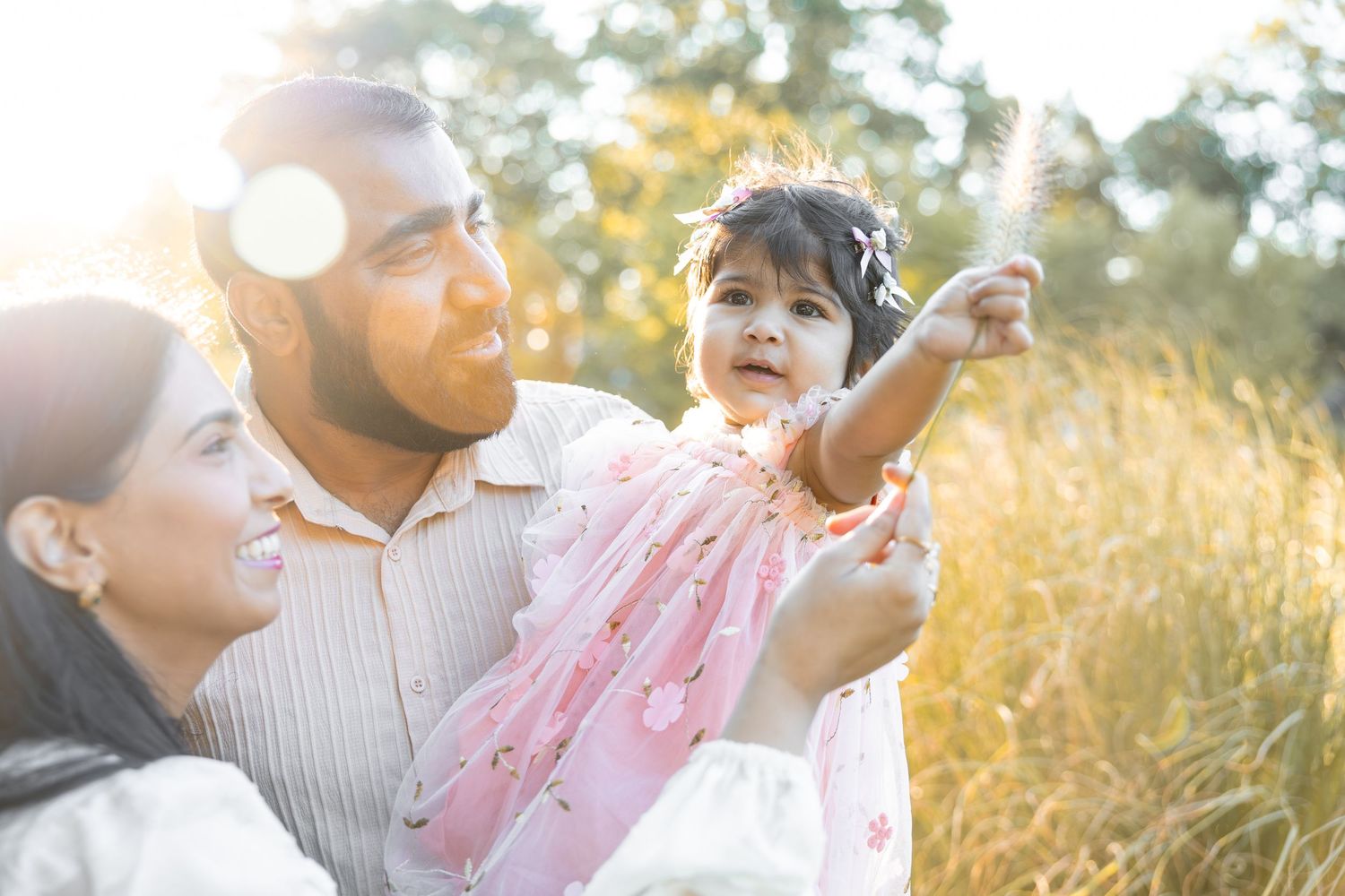 A smiling Asian family of three poses outdoors near a lake surrounded by lush green trees and grass.