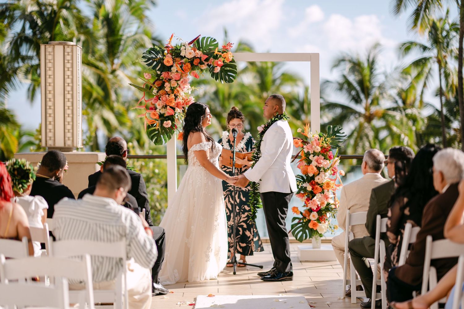 Couple exchanges vows at tropical outdoor wedding ceremony with floral arch and palm trees.