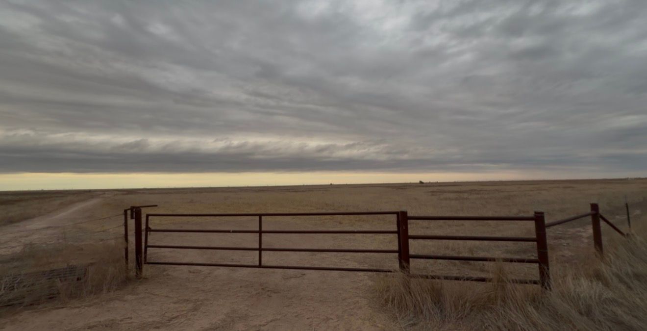 A rustic red metal farm gate on a dirt road overlooking vast open plains under a dramatic cloudy sky.