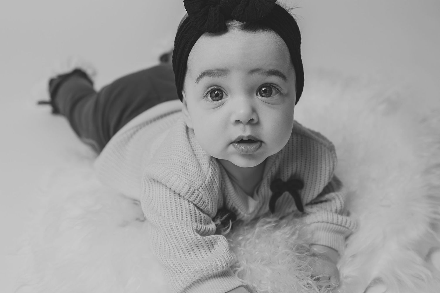 Black and white photo of an adorable baby lying on fluffy fur, wearing a knit sweater and hair bow.