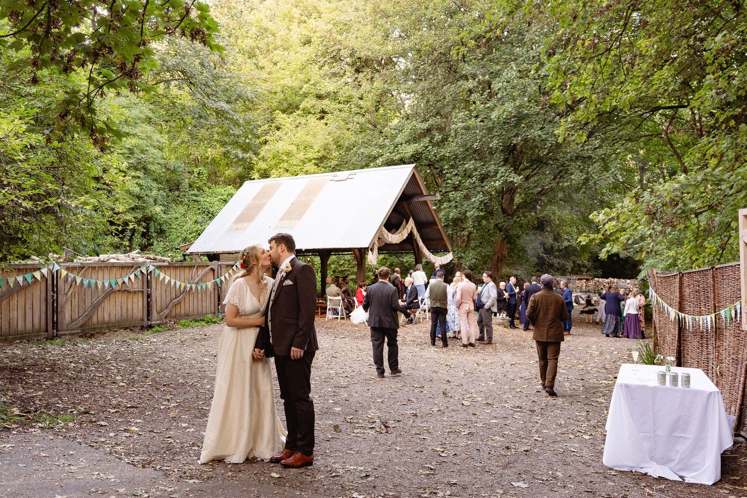 Bride and groom share a kiss at an outdoor woodland wedding venue with guests mingling in the background.