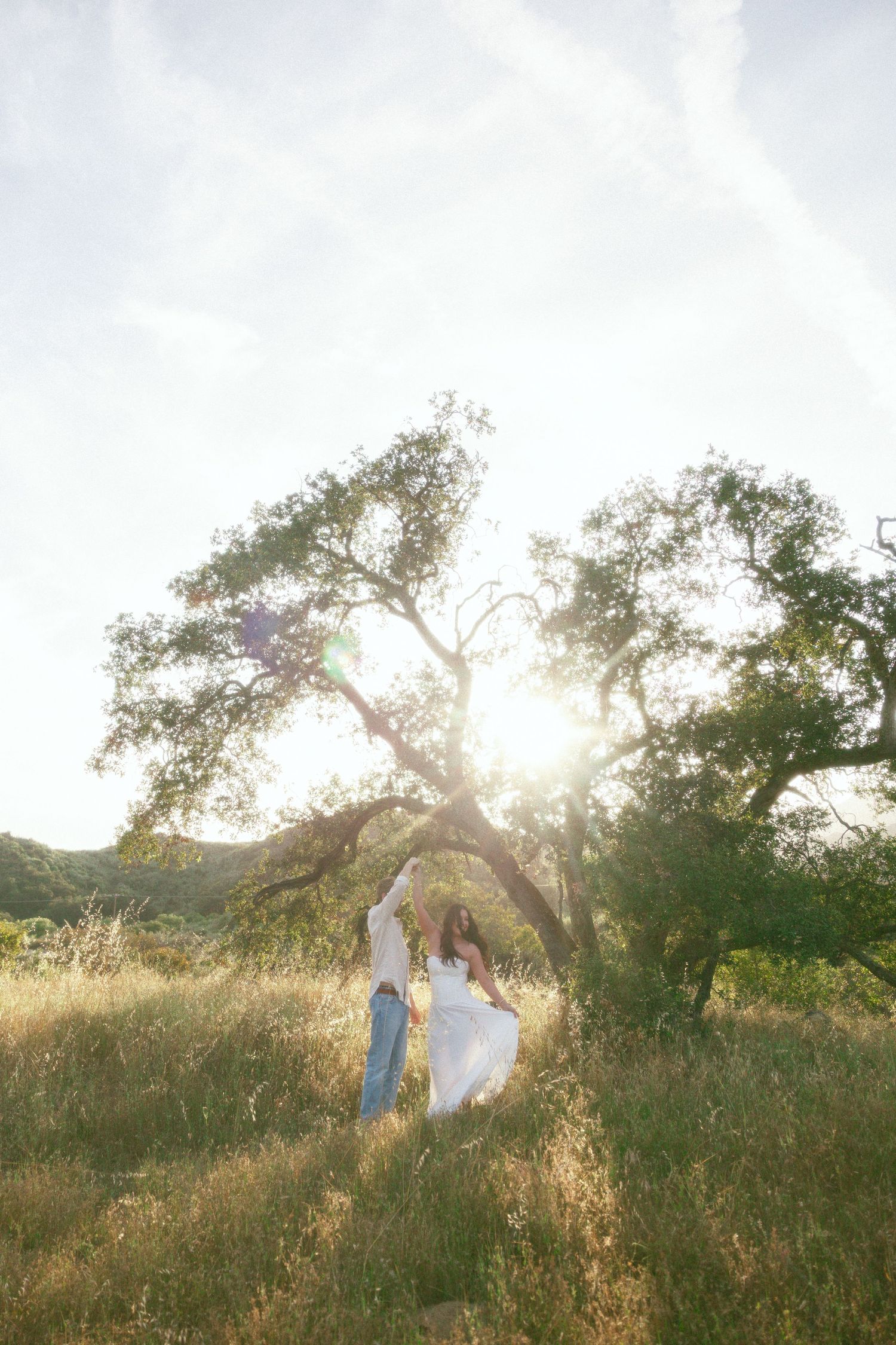 Couple embracing in a wildflower meadow with mountain backdrop, woman in white dress, man in casual linen shirt.