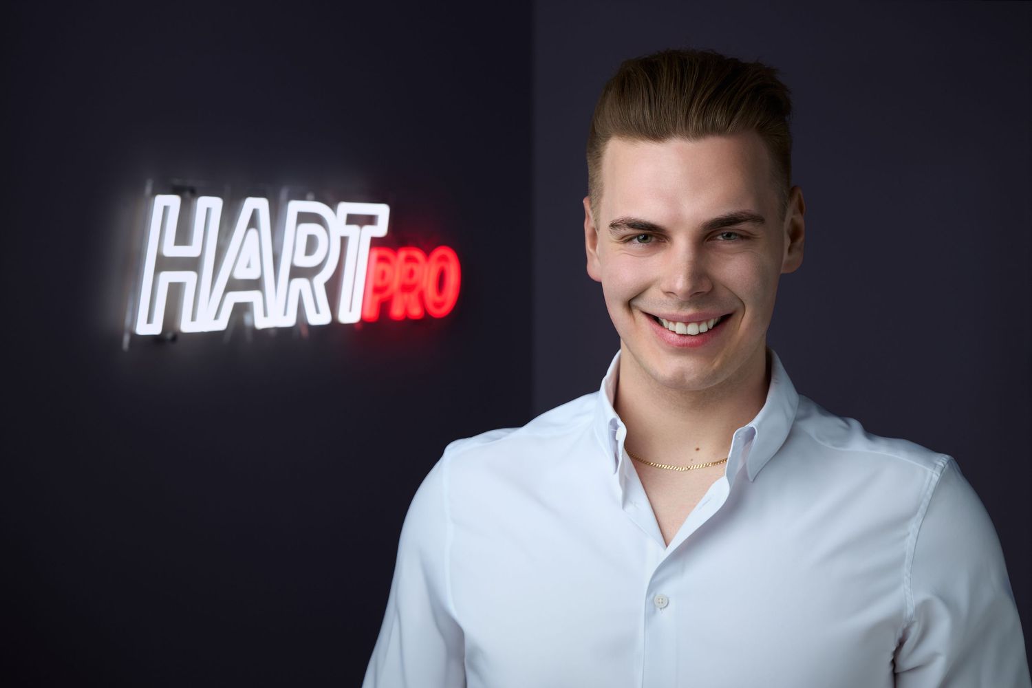 Young man smiling in front of a HartPro neon sign logo on a dark background, wearing a white dress shirt.