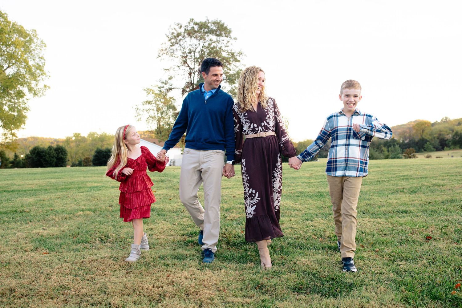 Family of four holding hands walking in a green field during golden hour, wearing coordinated outfits.