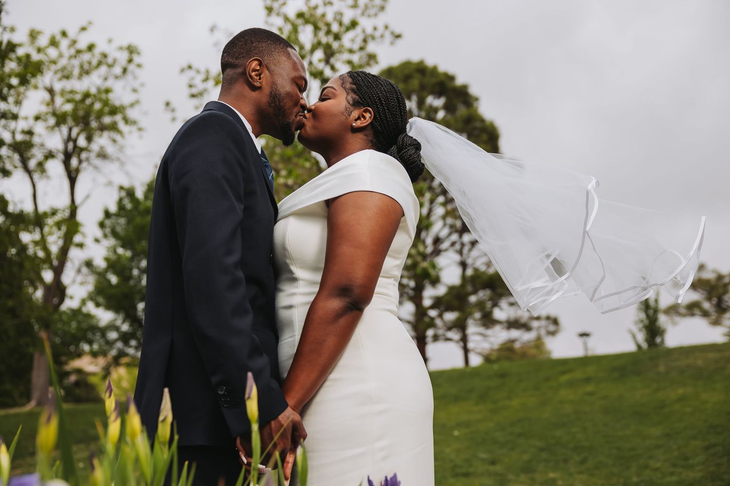 A bride and groom share a romantic kiss outdoors on their wedding day, with the bride's veil blowing in the wind.