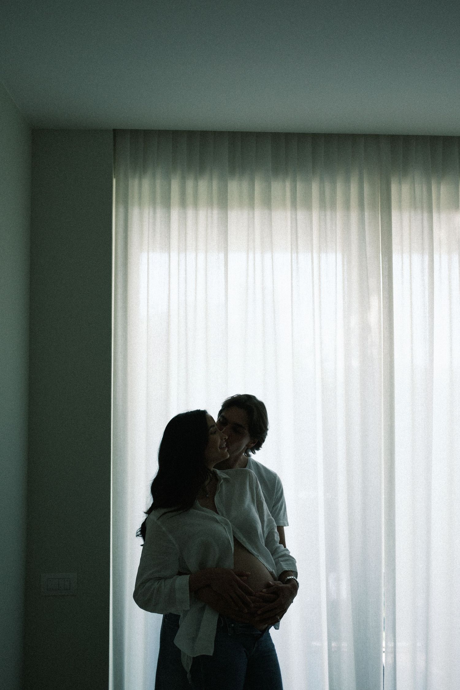 Couple embracing romantically in silhouette against sheer white curtains in a softly lit room.