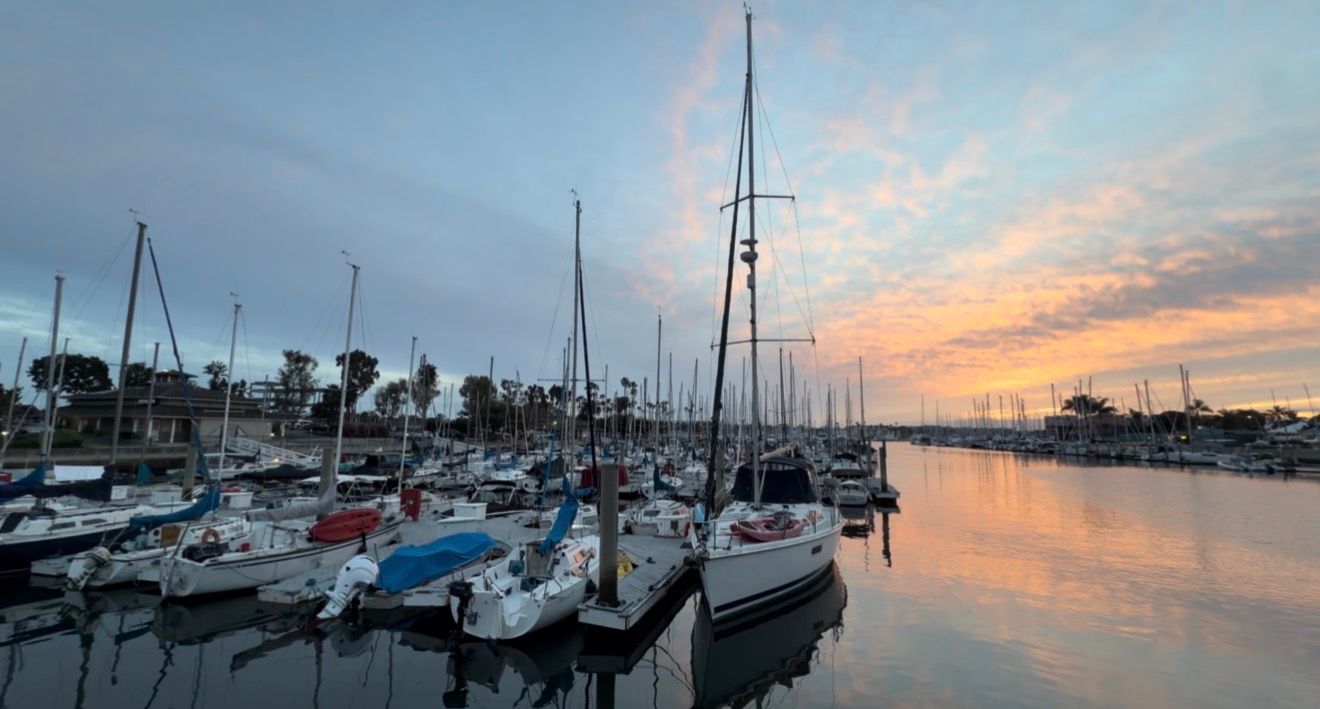 A panoramic view of boats docked at a marina during a colorful sunset with orange and pink clouds reflecting on calm waters.