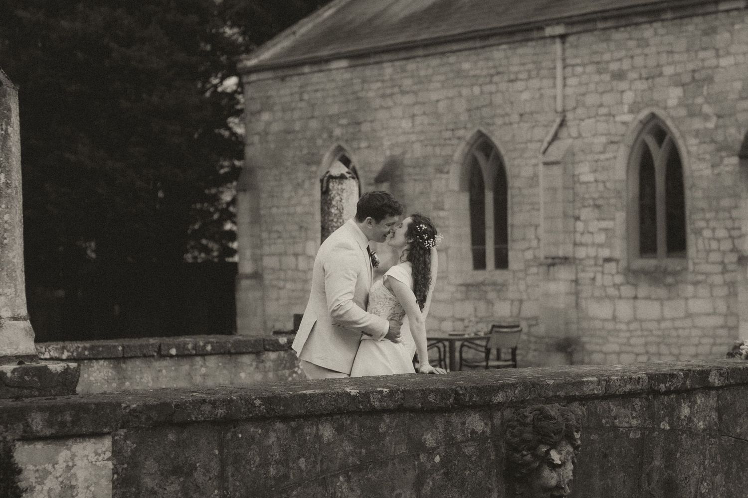 Black and white photo of a couple sharing a romantic kiss near a stone church wall on their wedding day.