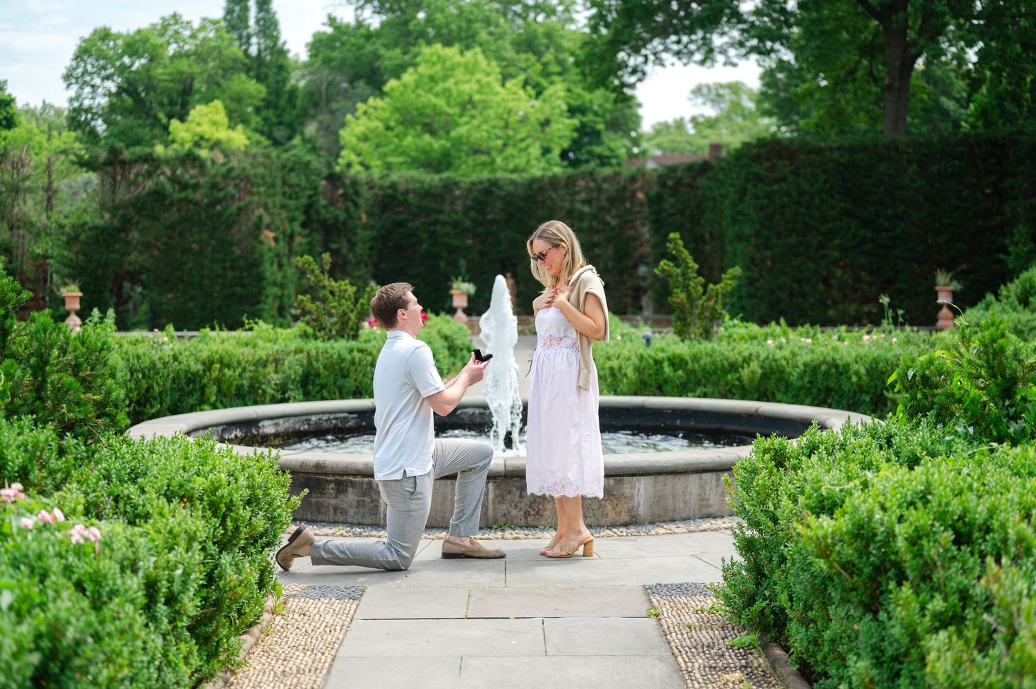 Man proposes to woman on one knee beside a garden fountain surrounded by lush green hedges on a sunny day.