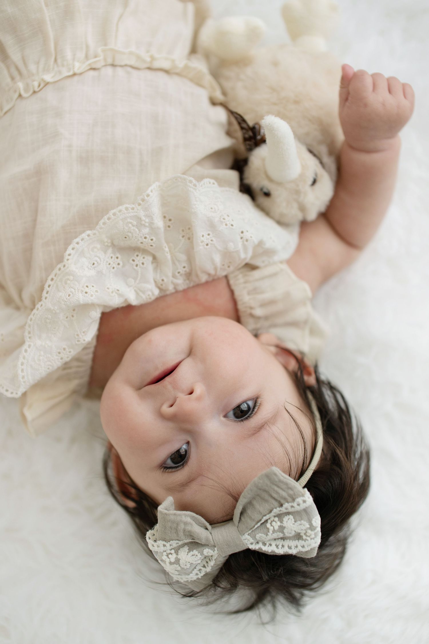 Baby girl in cream eyelet dress with gray bow headband lies on white fur, holding a plush bunny toy.