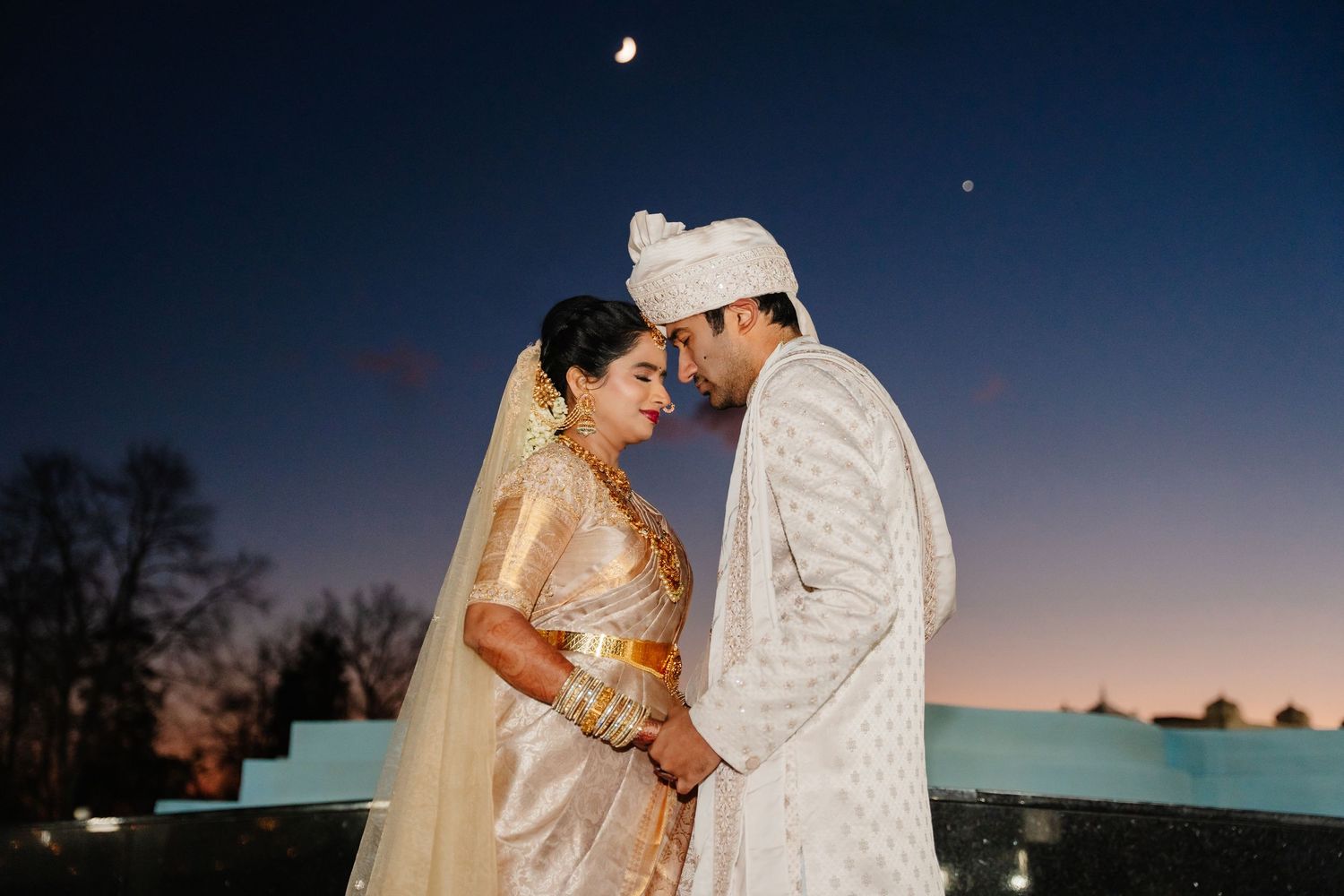 Indian bride and groom in traditional wedding attire share a romantic moment under a twilight sky.