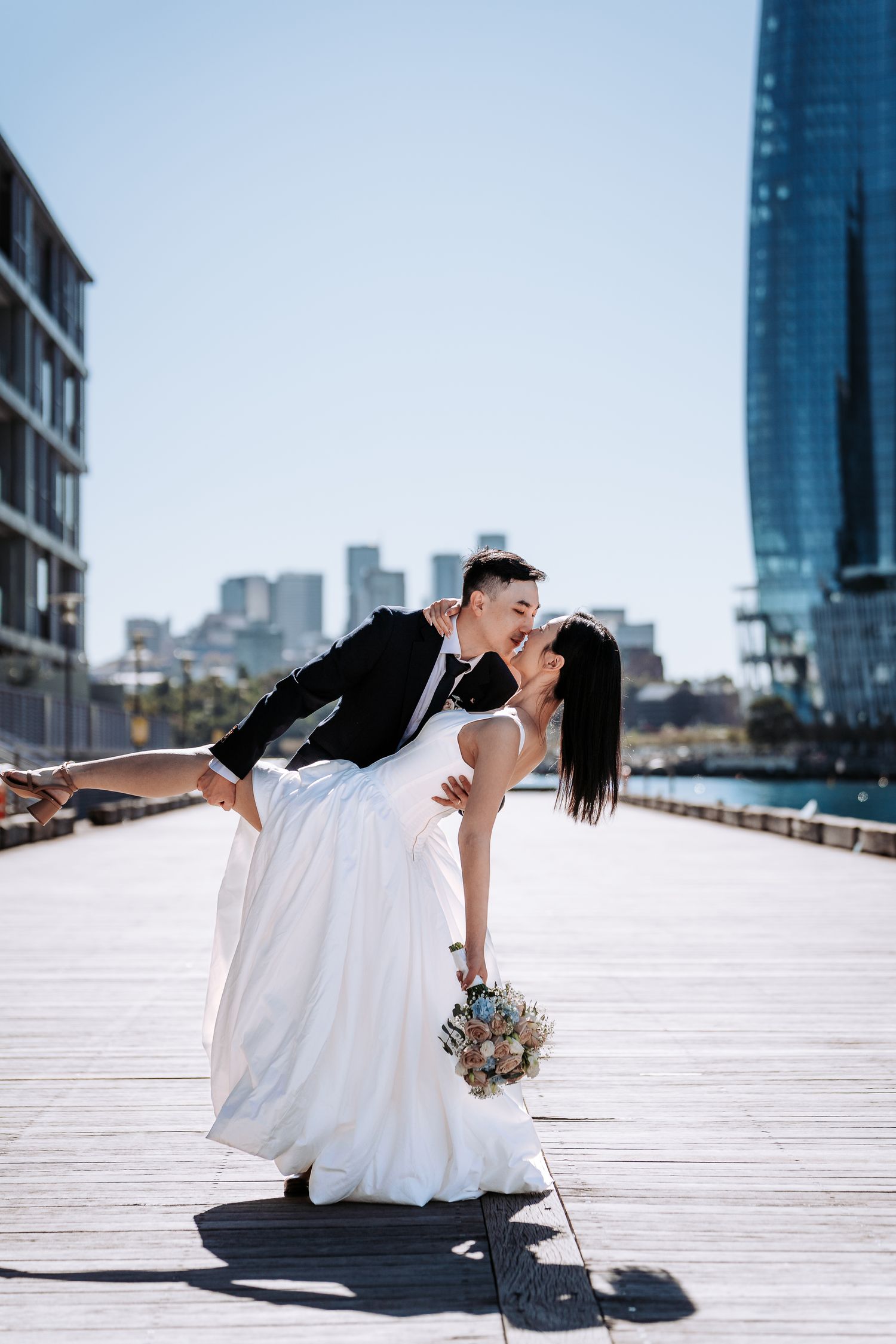 Bride lifts groom in a romantic dip kiss on a sunny urban boardwalk, holding a bouquet with city skyline behind them.