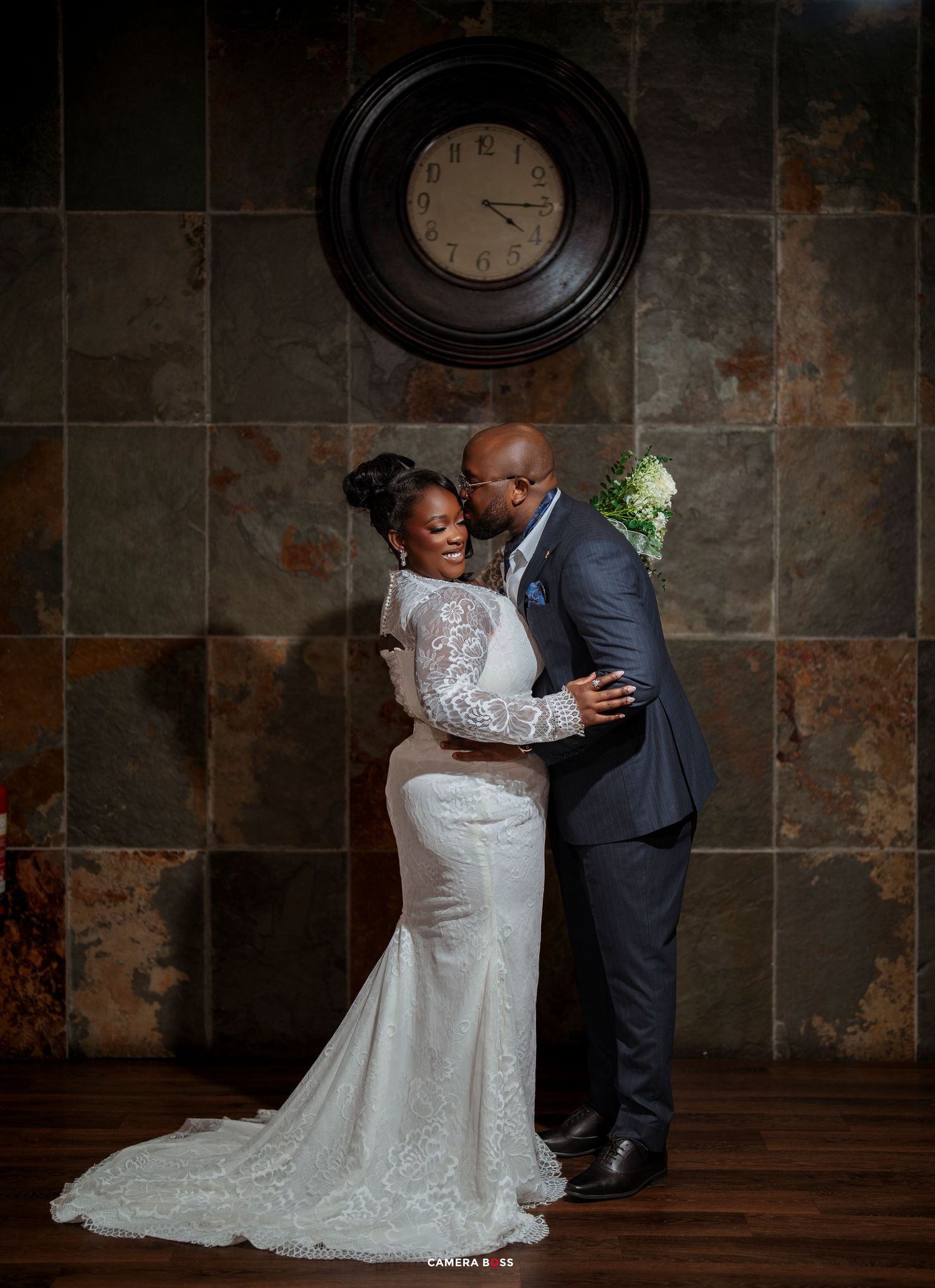 Bride and groom share a romantic embrace in wedding attire before a rustic stone wall with a vintage clock.