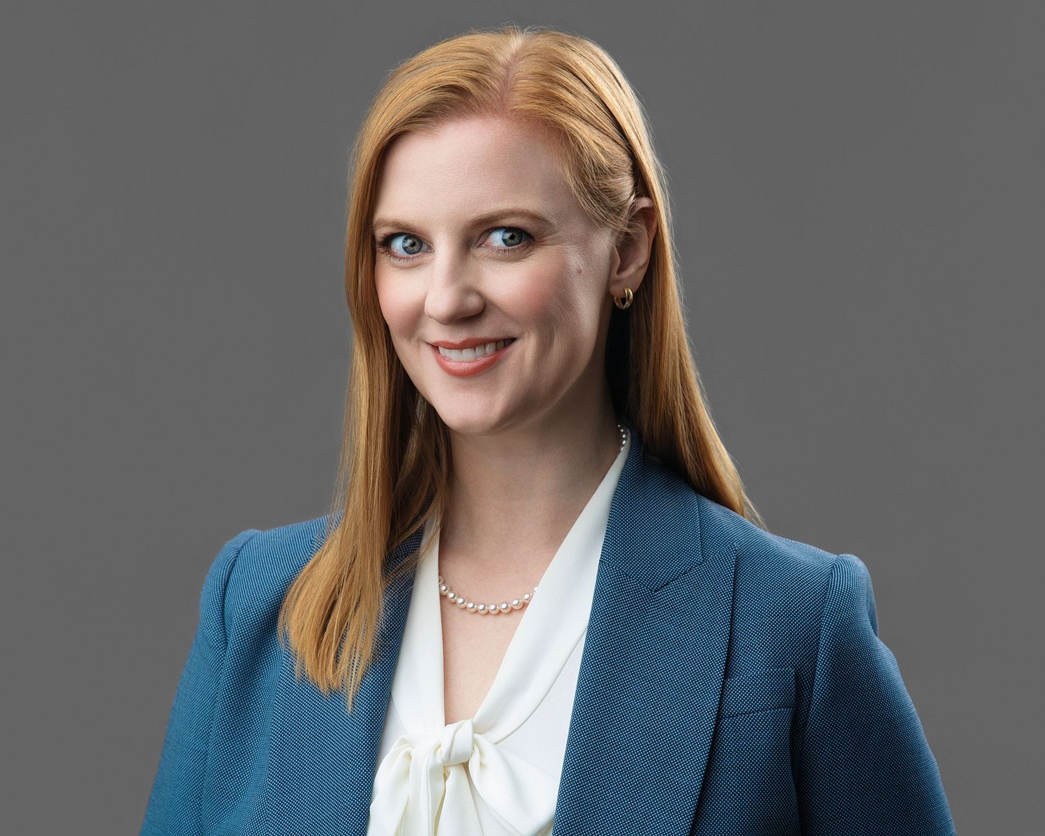 Professional woman with red hair in a teal blazer and white blouse, smiling against a gray background.